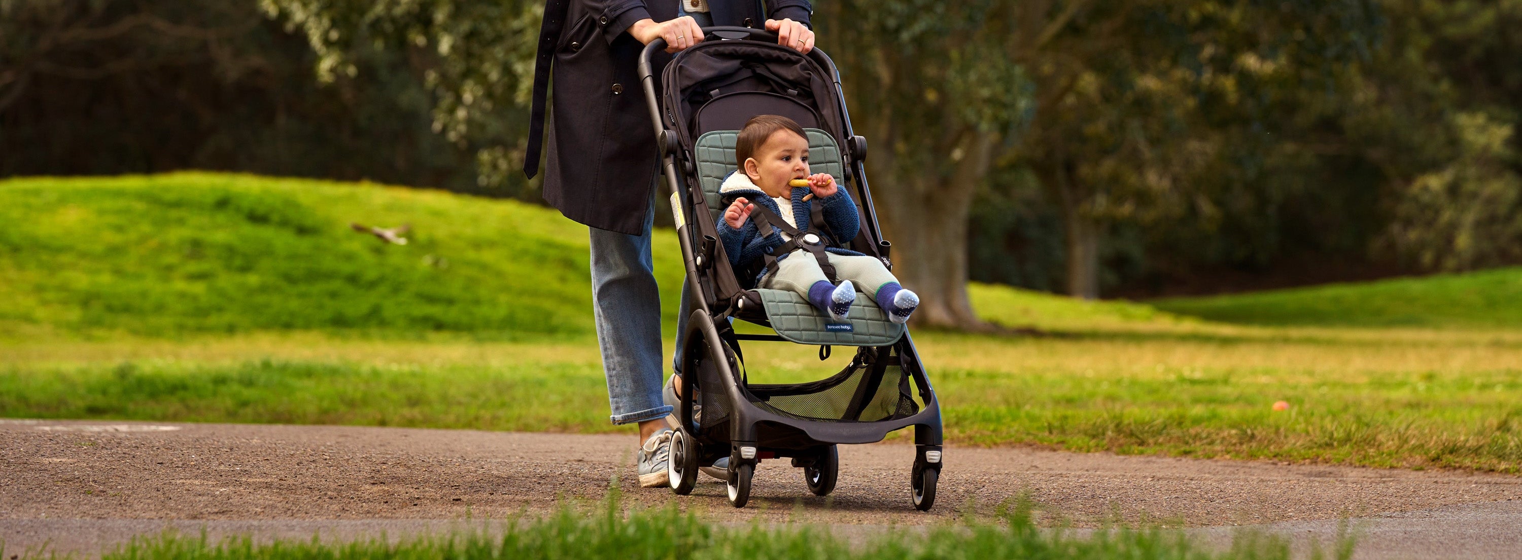 A woman in a dark coat and jeans pushes a stroller with a baby inside along a paved path in a grassy park with trees in the background.