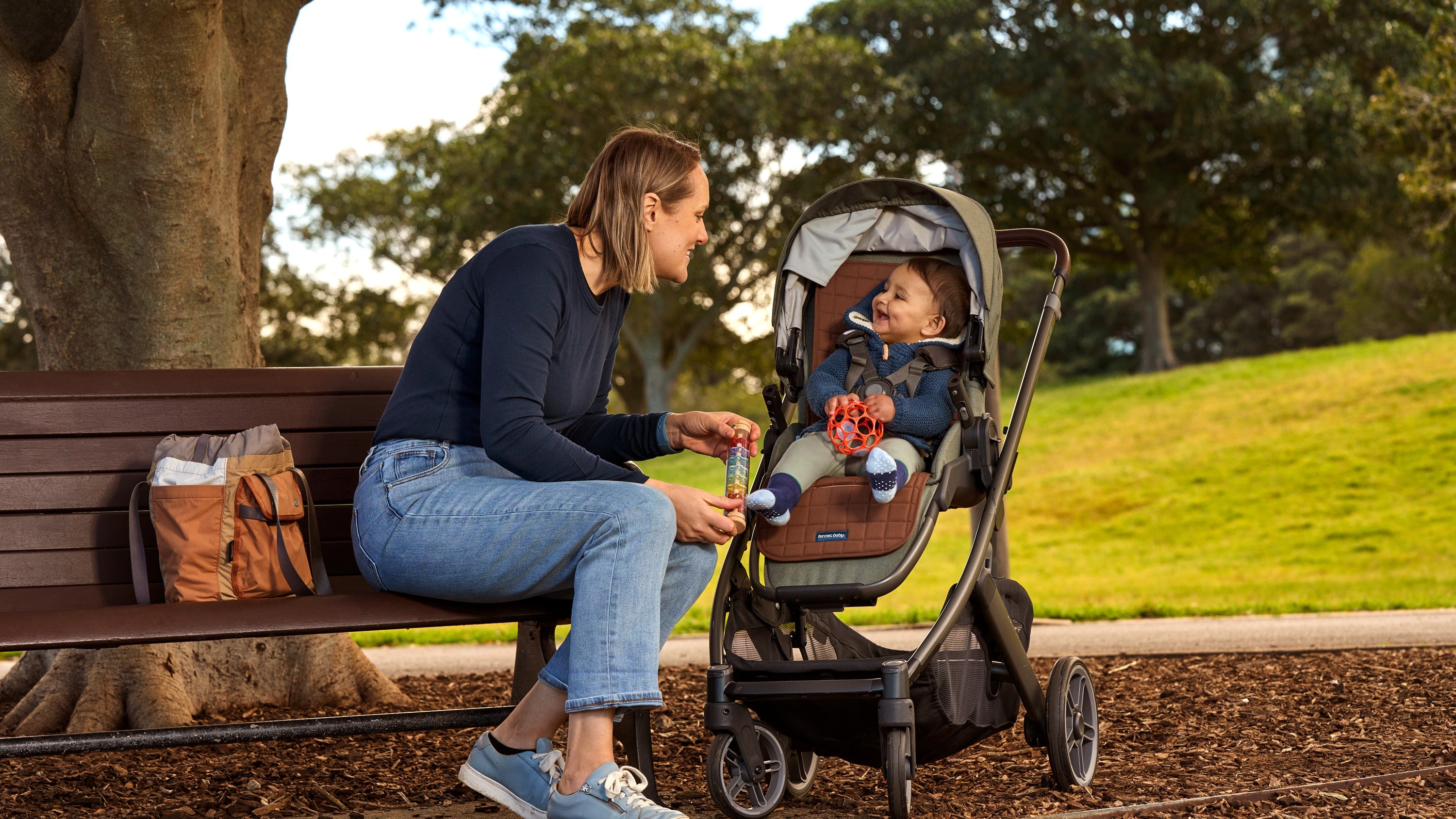 A woman sits on a park bench next to a stroller with a child inside, outdoors on a mulch path with grass and trees in the background.